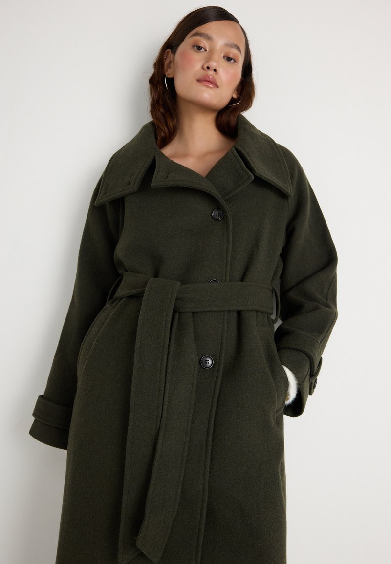 Mulher vestindo um longo casaco de lã verde escuro, com cinto, colarinho largo e botões, de pé contra um fundo branco liso.