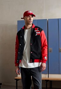 Giacca varsity nera e rossa con bottoni, caratterizzata da toppe e un logo, indossata sopra una maglietta bianca. Accompagnata da un cappellino rosso.