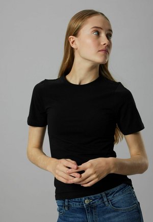 Young woman with long light hair wearing a black short-sleeve top and blue jeans, looking to her right, hands loosely clasped.