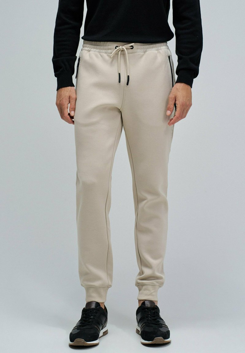 Hombre vestido con joggers beige con bolsillos con cremallera, zapatillas negras y una camiseta de manga larga negra, de pie frente a un fondo gris claro.