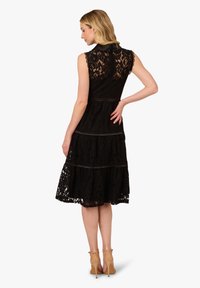 Abito in pizzo nero con design a strati, senza maniche e con un collo alto. Presenta dettagli in pizzo intricati e una chiusura con zip sul retro.