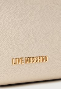 Υφή από μπεζ δέρμα με ανάγλυφο σχέδιο και μεταλλικό χρυσό λογότυπο "LOVE MOSCHINO" στη μέση κοντά στο κάτω άκρο.