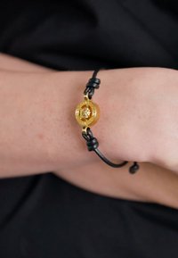 Bracciale in pelle nera con un accento circolare dorato caratterizzato da una superficie testurizzata e un centro dorato, fissato con cordini regolabili.