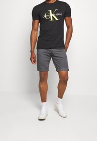 Mężczyzna ubrany w czarną koszulkę Calvin Klein Jeans, szare, podwinięte spodenki, białe skarpetki i białe sneakersy, stoi na jednolitym jasnym tle.