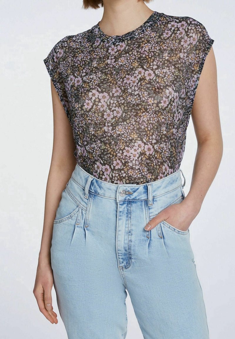 Femme portant un haut transparent noir à motifs floraux, rentré dans un jean bleu clair taille haute, avec une main dans la poche, sur un fond uni.