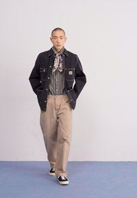 Black denim jacket, striped button-up shirt, tan trousers, patterned scarf, and black sneakers. Model stands on a blue floor against a white background.
