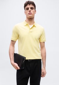 Νέος άνδρας που φοράει κίτρινο πόλο πουκάμισο και μαύρο παντελόνι, κρατώντας μια μαύρη θήκη με φερμουάρ στο αριστερό του χέρι.