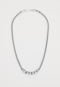 Collier en métal argenté avec une chaîne tressée, comportant des blocs de lettres formant le mot "LACOSTE" dans une police en gras et carrée.