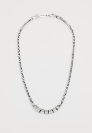 Collana in metallo argentato con catena intrecciata, caratterizzata da blocchi di lettere che compongono "LACOSTE" in un carattere audace e squadrato.