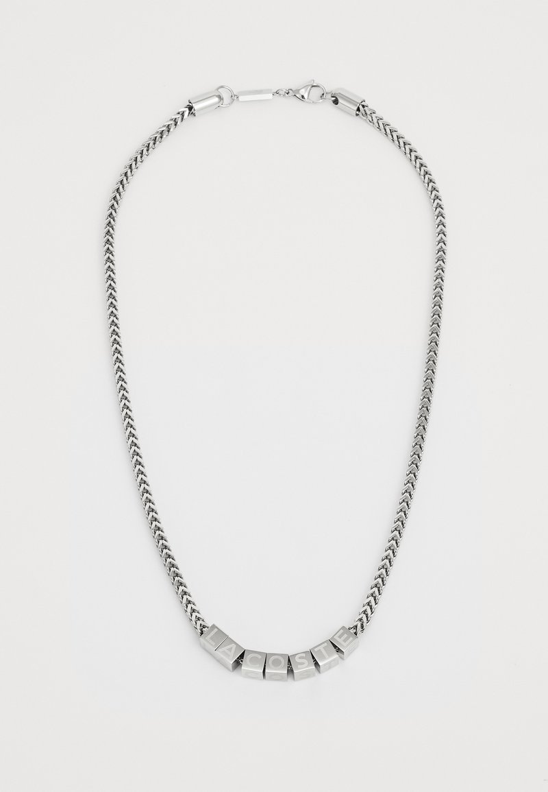 Collier en métal argenté avec une chaîne tressée, comportant des blocs de lettres formant le mot "LACOSTE" dans une police en gras et carrée.