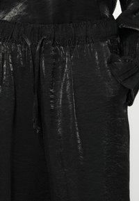 Čierne textúrované nohavice s lesklým povrchom, elastickým pásom s šnúrkou a bočnými vreckami; tkanina sa javí ako ľahká a jemná.