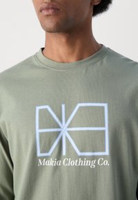 Olíva zöld hosszú ujjú póló kék geometrikus logóval és "Makia Clothing Co." felirattal, puha pamut anyagból készült.