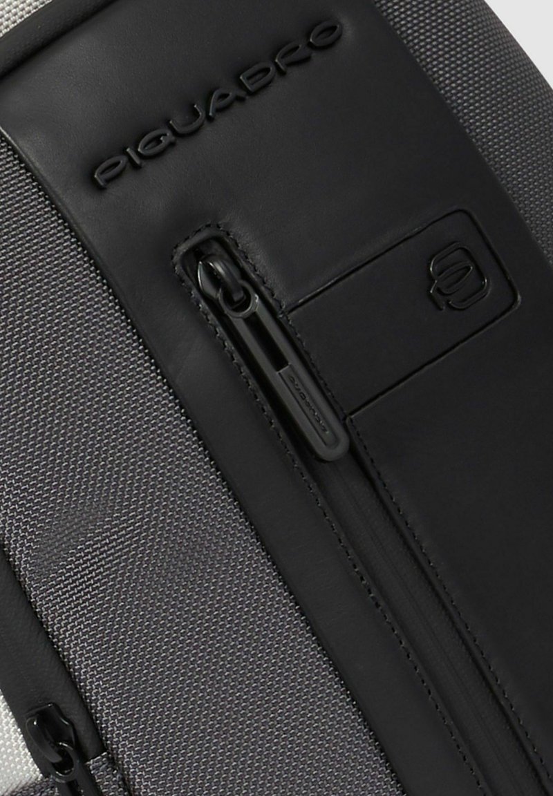 Primo piano di una sezione di borsa in tessuto grigio e pelle nera con una cerniera verticale e il nome del marchio "Piquadro" in rilievo sulla pelle.