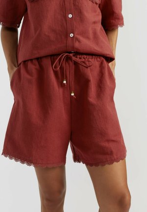 Femme portant un short couleur rouille avec cordon et bordure en dentelle, et une chemise boutonnée assortie avec manches en dentelle, mains dans les poches.