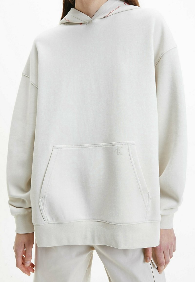 Sudadera color crema con un bolsillo frontal tipo canguro, puños y dobladillo acanalados, que presenta un logo en el bolsillo y un detalle estampado en la capucha.
