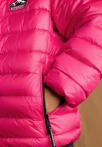 Jasne ružové prešívané búcha s horizontálnym prešívaním, s vreckom na zips a nášivkou loga Superdry na pleci. Mäkka, polstrovaná textúra.