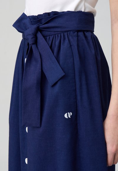 Jupe bleu marine avec une taille nouée, en tissu texturé, boutons sur le côté et un petit logo blanc près de l'ourlet.
