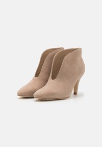 Un paio di stivaletti beige in suede con punta affilata, taglio frontale a V e tacchi a spillo di altezza media su sfondo bianco.