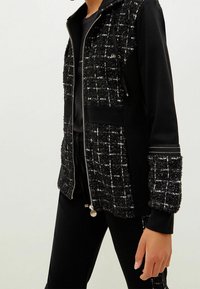 Chaqueta negra con cremallera que presenta un frente de tweed texturizado con un patrón de cuadros, mangas negras sólidas y acentos en hardware plateado.