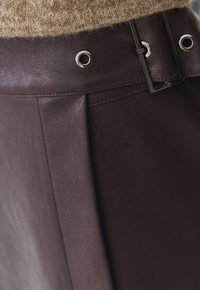 Gonna in pelle marrone con design a pieghe, vita con occhielli e fibbia metallica. Texture liscia con una leggera lucentezza.