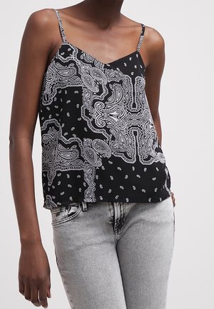 Nainen, joka pukeutunut mustavalkoiseen paisley-kuvioiseen hihattomaan toppiin, jossa ohuet olkaimet, ja vaaleanharmaisiin korkeavyötäröisiin farkkuihin.