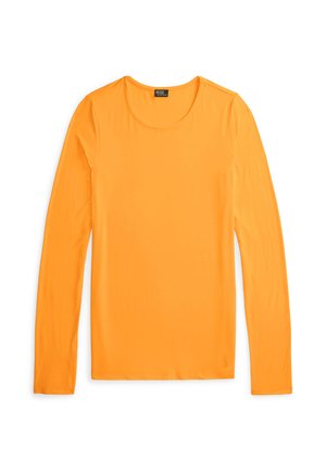Modelka nosząca pomarańczową bluzkę z długim rękawem oraz jasne zielone, szerokie, w paski spodnie z widocznym paskiem Ralph Lauren.
