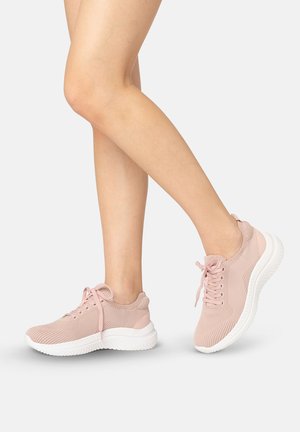 Vrouwelijke benen met lichtroze gebreide sneakers met witte zolen, één voet plat en de andere op de tenen tegen een grijze achtergrond.