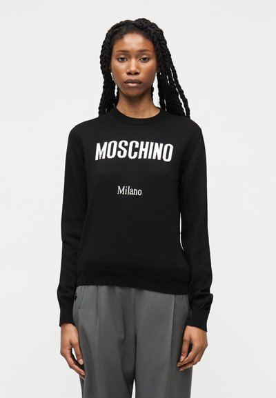 Femme aux longues tresses portant un sweat-shirt noir avec le texte "Moschino Milano" et un pantalon gris, sur un fond clair uni.
