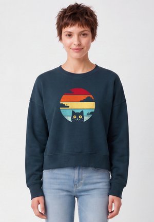 Jeune personne aux cheveux courts portant un sweat-shirt bleu foncé avec un coucher de soleil coloré et un graphique de chat noir, associé à un jean bleu clair.
