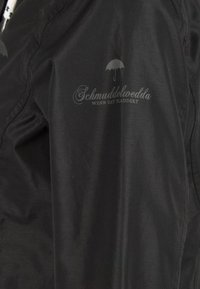 Chaqueta negra hecha de un material suave, con un logotipo sutil y un gráfico de paraguas en el pecho. Tiene un diseño moderno y una textura ligera.