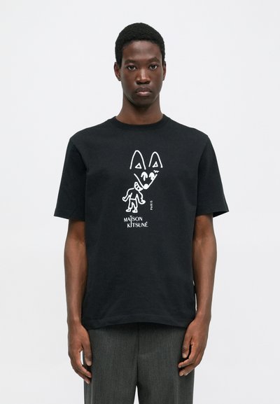 Muž v černém tričku Maison Kitsuné s bílým abstraktním grafikem lišky a postavy, stojící na jednobarevném bílém pozadí.