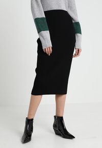 Jupe midi noire à côtes, associée à un pull gris avec des rayures vertes en accent. Le mannequin porte des bottines noires brillantes à bout pointu.