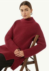 Burgundowy sweter prążkowany z wysokim kołnierzem, szerokimi rękawami i bocznymi rozcięciami, noszony na czarnych spodniach, siedząc na drewnianym krześle.