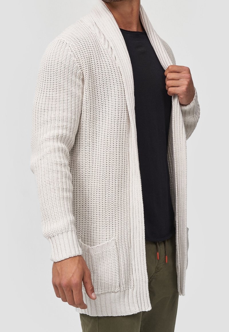 Homme portant un cardigan en tricot beige sur une chemise noire et un pantalon vert olive, tenant le col du cardigan avec la main droite.