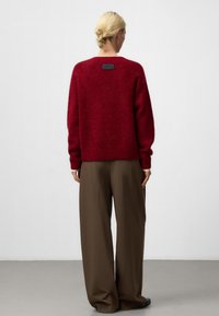 Czerwony sweter z wełny o luźnym kroju, z teksturowanym wykończeniem i czarną metką na plecach. Połączony z luźnymi brązowymi spodniami.