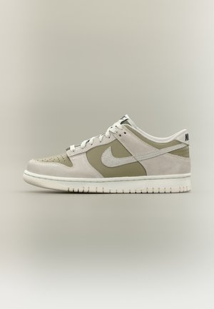 Baskets basses Nike en daim beige et blanc cassé avec des lacets blancs et une semelle blanche, vues de côté sur un fond neutre.