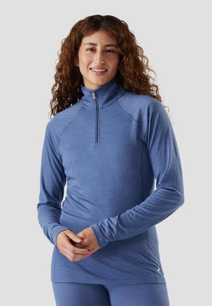 Mujer con cabello rizado que lleva una camiseta azul de manga larga con cremallera y pantalones a juego, sonriendo, de pie con las manos entrelazadas.