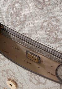 Beżowa torebka o strukturze z wytłoczonym wzorem logo, złotymi elementami i gładką podszewką wewnętrzną z nazwą marki w ciemniejszym kolorze.