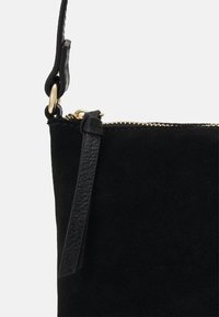 Borsa in suede nera con tracolla in pelle, zip dorata e hardware ad anello dorato su sfondo bianco.
