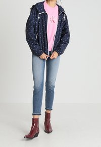 Giacca a pioggia blu navy con cappuccio, motivi bianchi, cerniera frontale, abbinata a una felpa rosa, jeans azzurri e stivaletti lucidi color bordeaux.