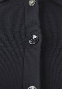 Zwarte ribgebreide cardigan met een close-up van een glanzende knop en een decoratieve bloemenaccent erboven. Focus op textuur en knoopdetail.