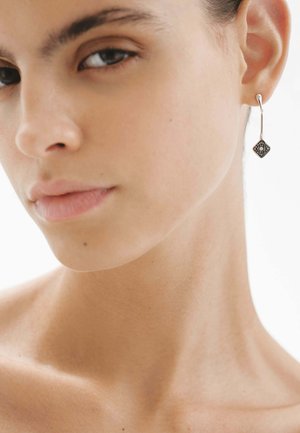 Pendiente de aro de plata con un colgante en forma de diamante que presenta un acento texturizado en negro y patrones circulares, suspendido de un gancho delgado.