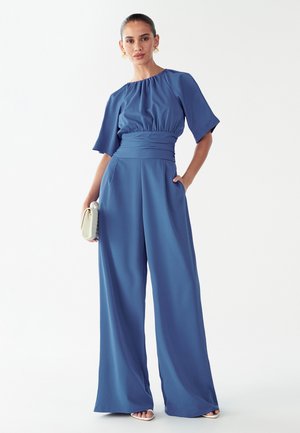 Blå jumpsuit med vide ben, samlet halsudskæring og korte ærmer. Glat stof med taljering i livet. Model holder en lille tekstureret clutch.
