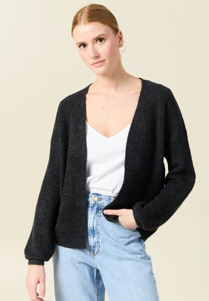 Schwarze, offene Strickjacke mit weichem Material, langen Ärmeln und lockerer Passform, kombiniert mit einem weißen V-Ausschnitt-Oberteil und hochgeschnittenen Jeans.