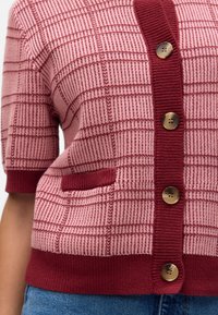 Gros plan d’un cardigan tricoté rose et bordeaux à motif à carreaux, quatre boutons, manches courtes, petite poche et bords côtelés sur un jean bleu.