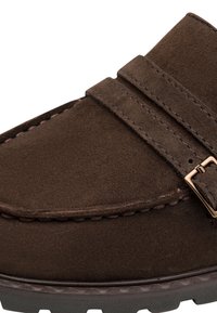 Mocassin en daim marron avec double bride et boucle dorée, coutures détaillées et semelle épaisse en caoutchouc.