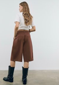 Shorts marroni a vita alta realizzati in un tessuto strutturato, con tasche posteriori, abbinati a stivali neri fino al ginocchio e una t-shirt bianca aderente.