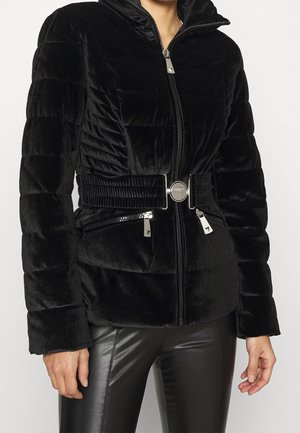 Schwarze gesteppte, samtige Jacke mit Frontreißverschluss, breitem Gürtel mit Metallschnalle, Reißverschlusstaschen, getragen über schwarzen Lederhosen.