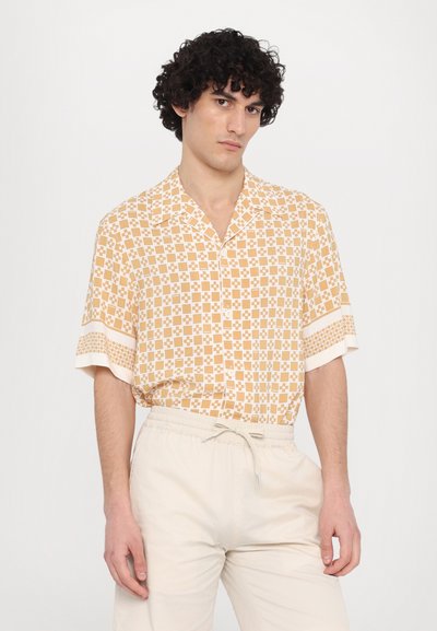 Chemise à manches courtes en orange clair avec un motif géométrique et des accents blancs, dotée d'un col et de boutons. Associée à un pantalon crème.