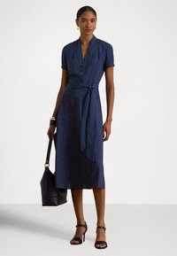 Kvinne iført en marineblå midi omslagskjole med korte ermer og hvite prikkete, svarte sandaler med hæl, og bærende en svart håndveske.
